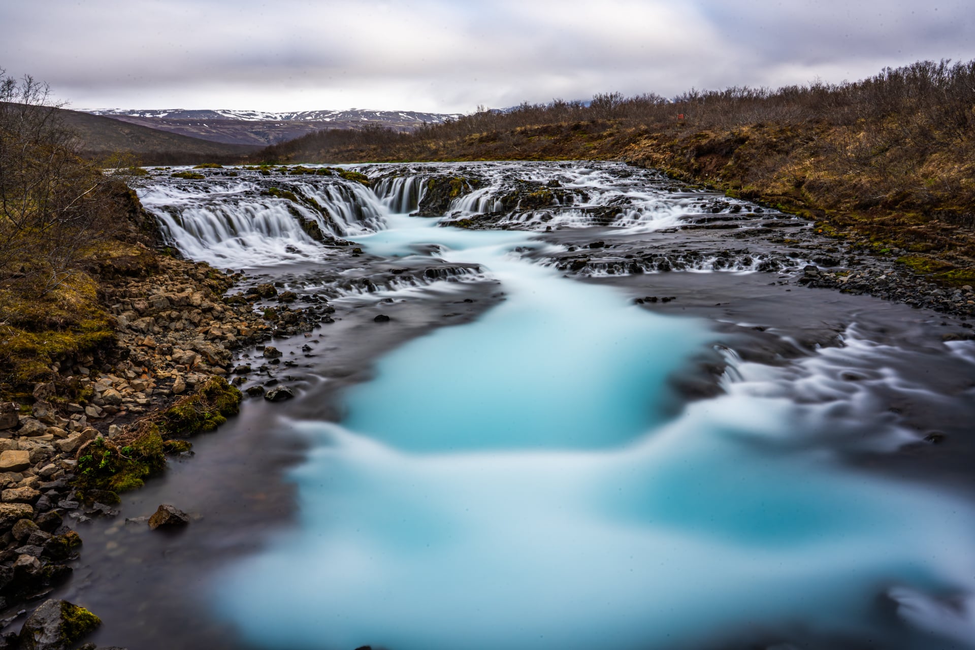 Long exposure photography of waterfall in Iceland. Water is almost azure blue with the sky being full overcast adding to the dramatic scenery.